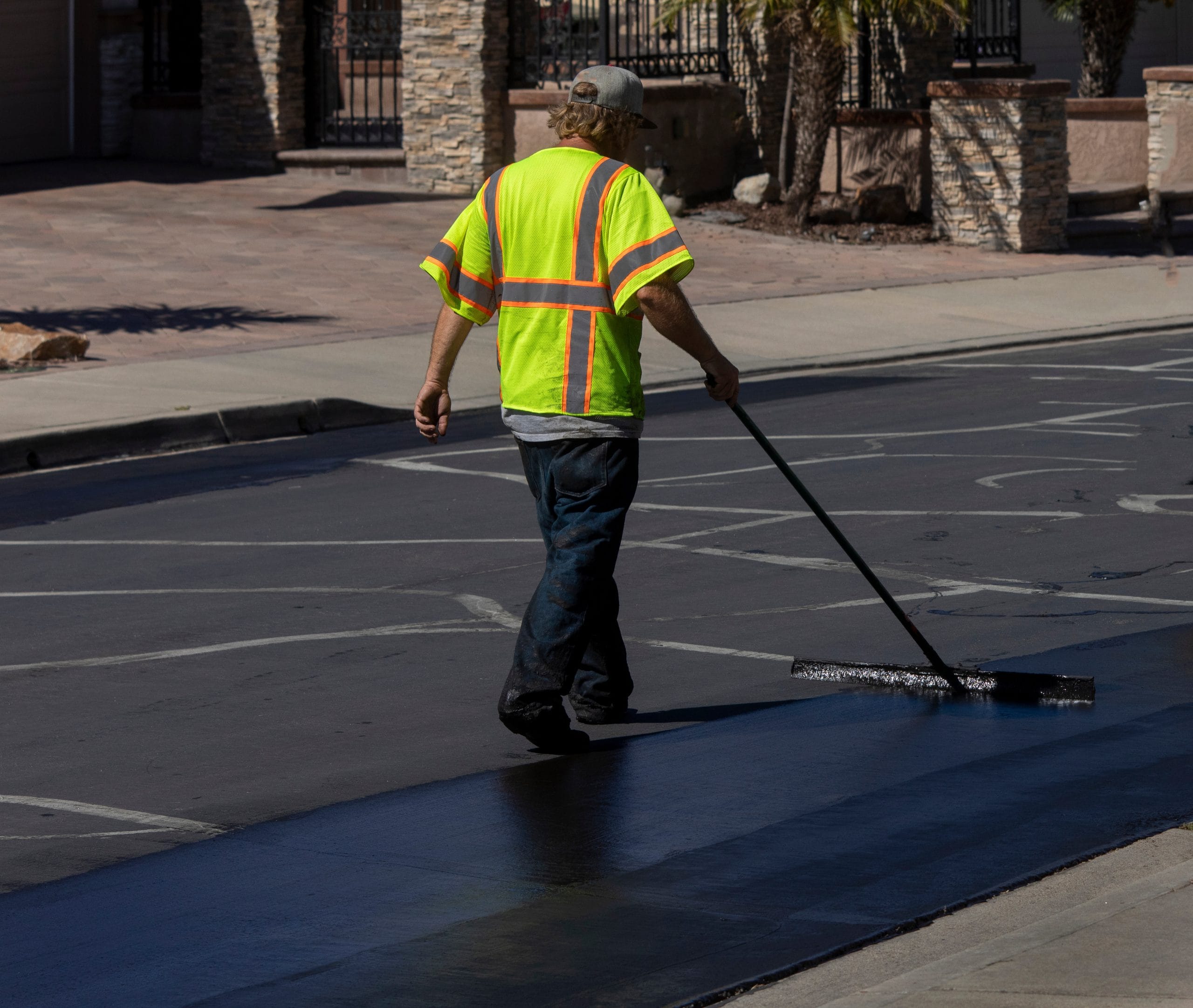 Worker using a sealcoating brush during asphalt resurfacing project Is Your Asphalt Aging Faster Than It Should? How Sealcoating Can Turn Back the Clock Worker in a neon yellow vest spreads asphalt on a street with a long-handled tool.