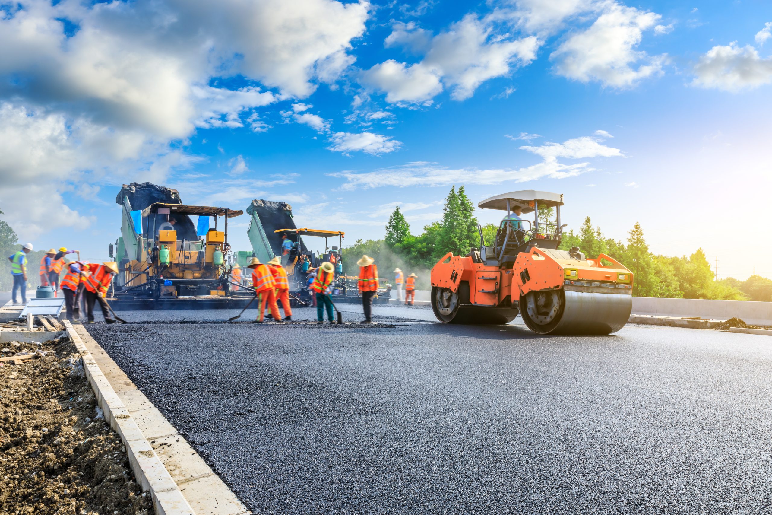 Construction workers paving a road with asphalt using large machines under a blue sky.