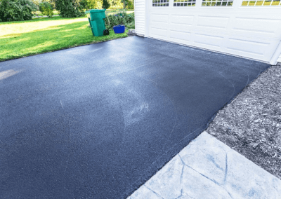 Freshly paved driveway leading to a white garage, with green lawn and trees in the background.