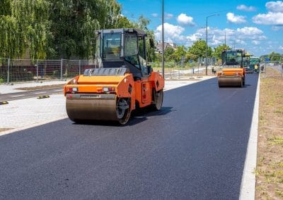 Two orange road rollers compress fresh asphalt on a street, with trees and blue sky in the background.