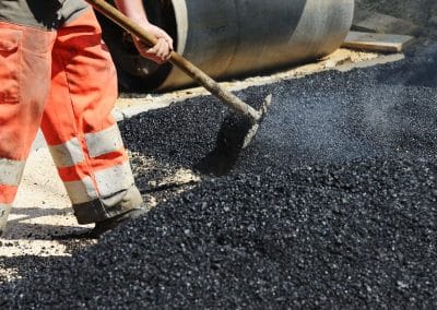 Worker in orange pants spreading asphalt with a rake on a construction site, with a steamroller in the background.