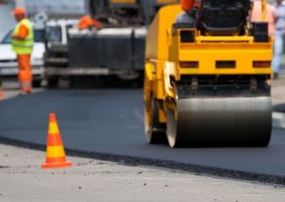 Construction workers in orange uniforms using machinery to lay asphalt on a road, with a traffic cone nearby.