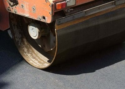 Close-up of an orange asphalt roller smoothing newly laid black pavement in bright sunlight.