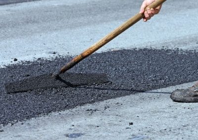 A person levels wet asphalt on a road with a wooden tool on a sunny day.