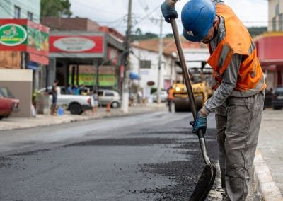 Worker in orange vest and blue helmet spreads asphalt on a road with a shovel in a busy town area.