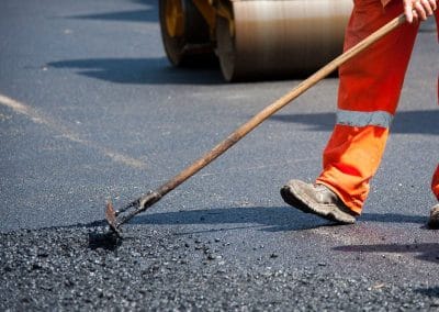 A worker in orange safety gear rakes fresh asphalt on a road, with a steamroller visible in the background.