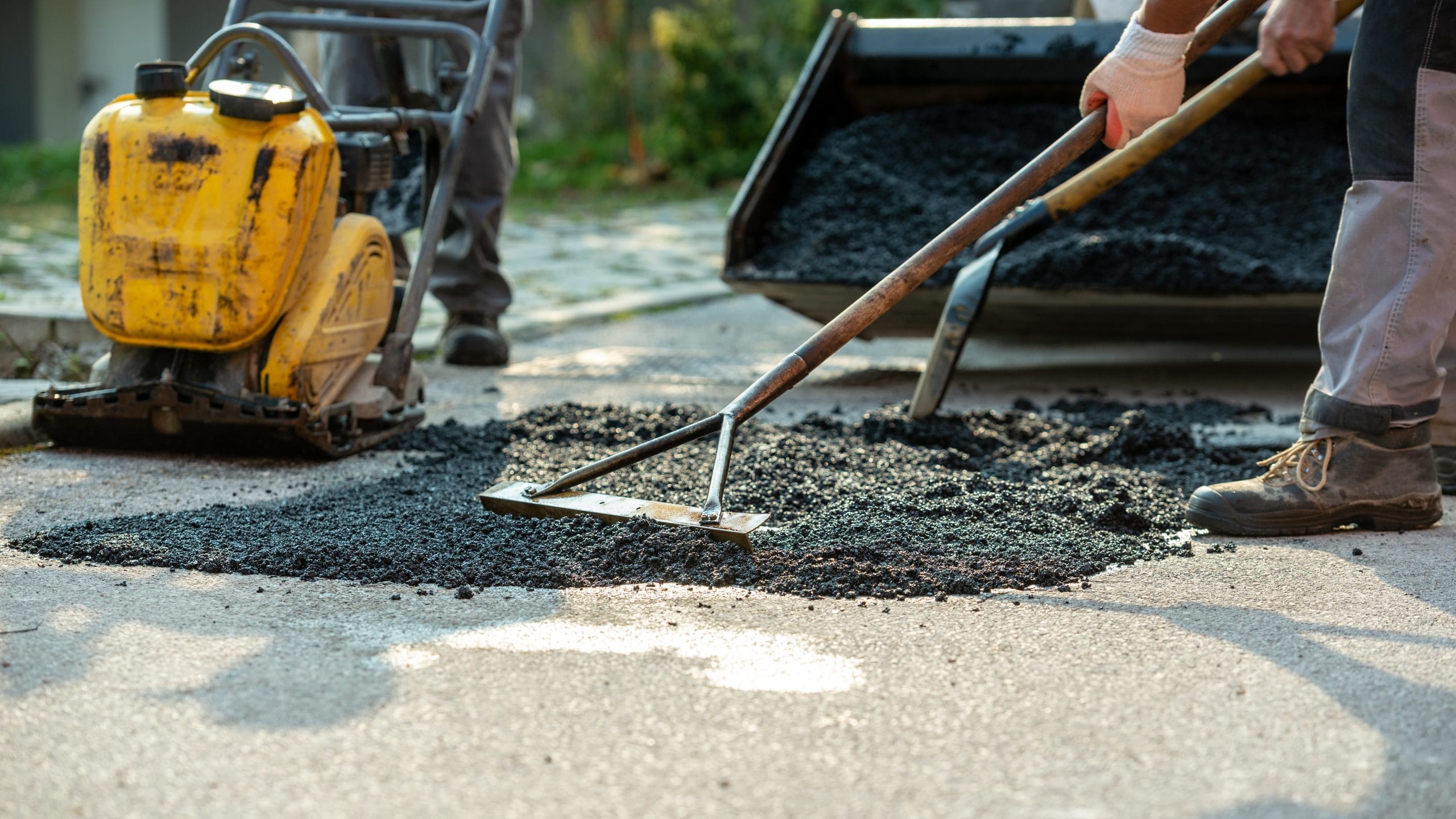 Workers raking fresh asphalt on a road, with a compactor machine nearby.