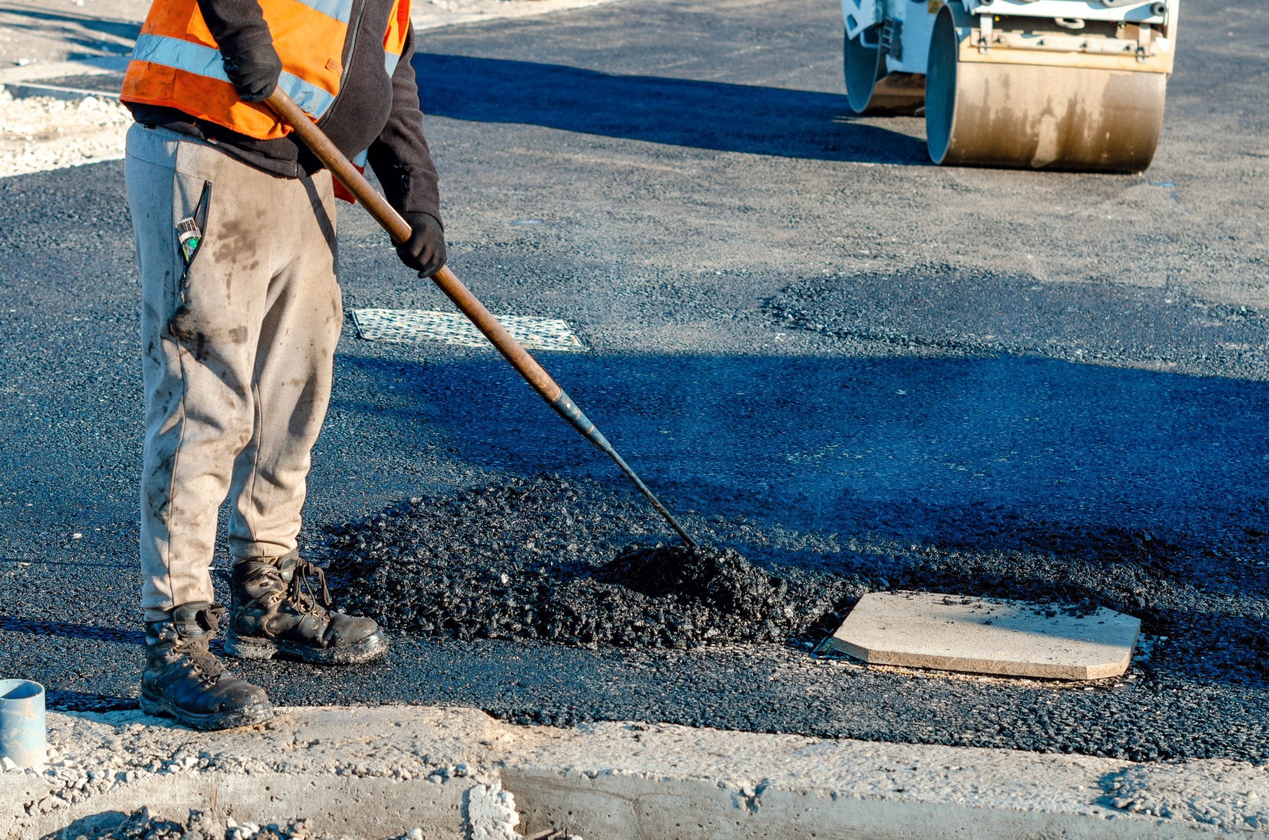Construction worker spreading asphalt on a road with a tool, wearing a safety vest and gloves.